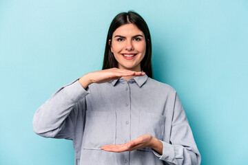 Young caucasian woman isolated on blue background holding something with both hands, product presentation.