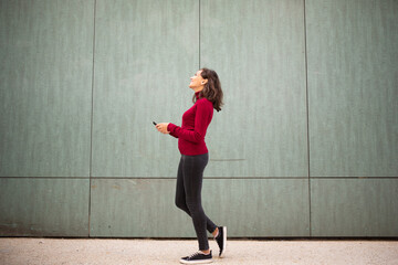 Full body young woman walking with cellphone by wall