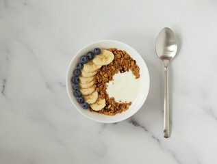 Bowl with granola, yougurt, banana and blueberry, spoon, marble background, flat lay