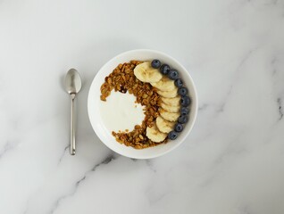Bowl with granola, yougurt, banana and blueberry, spoon, marble background, flat lay