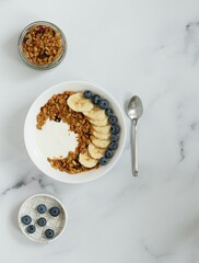 Bowl with granola, yougurt, banana and blueberry, jar with granola, saucer with blueberry, marble background, flat lay