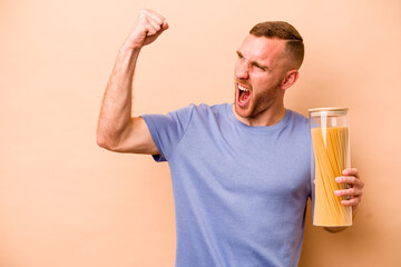 Young caucasian man holding spaghettis jar isolated on beige background raising fist after a victory, winner concept.