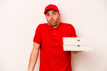 Young delivery caucasian man holding pizzas isolated on white background shrugs shoulders and open eyes confused.