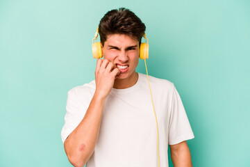 Young caucasian man listening to music isolated on blue background biting fingernails, nervous and...