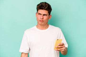 Young caucasian man holding mobile phone isolated on blue background confused, feels doubtful and unsure.