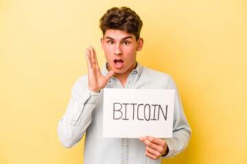 Young caucasian man holding bitcoin placard isolated on yellow background surprised and shocked.