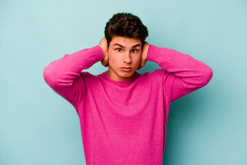 Young caucasian man isolated on blue background covering ears with hands trying not to hear too loud sound.