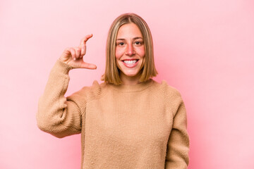 Young caucasian woman isolated on pink background holding something little with forefingers, smiling and confident.