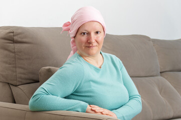 Mid adult woman with cancer in pink headscarf smiling sitting on couch at home. Smiling woman suffering from cancer sitting after taking chemotherapy sessions.