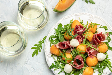 mediterranean salad with prosciutto or jamon, mozzarella and green basil leaves and Cantaloupe melon on white table, traditional Spanish and Italian appetizer served with wholemeal grissini, top view