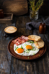 Fried eggs with bacon and herbs, 2 white bread toasts. On the table is a napkin, spices, a glass of champagne. Dark brown wooden background. Breakfast menu for cafe, restaurant, bar.
