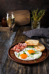 Fried eggs with bacon and herbs, 2 white bread toasts. On the table is a napkin, spices, a glass of champagne. Dark brown wooden background. Breakfast menu for cafe, restaurant, bar.