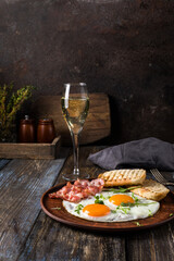 Fried eggs with bacon and herbs, 2 white bread toasts. On the table is a napkin, spices, a glass of champagne. Dark brown wooden background. Breakfast menu for cafe, restaurant, bar.