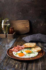 Fried eggs with bacon and herbs, 2 white bread toasts. On the table is a napkin, spices, a glass of champagne. Dark brown wooden background. Breakfast menu for cafe, restaurant, bar.