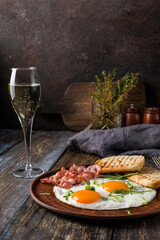 Fried eggs with bacon and herbs, 2 white bread toasts. On the table is a napkin, spices, a glass of champagne. Dark brown wooden background. Breakfast menu for cafe, restaurant, bar.