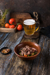 Deep plate with hot vegetables and beef. On a wooden table is a mug of light beer, spices and tomatoes. Menu for bar, cafe, restaurant. Dish from the chef. Close up