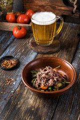 Deep plate with hot vegetables and beef. On a wooden table is a mug of light beer, spices and tomatoes. Menu for bar, cafe, restaurant. Dish from the chef. Close up