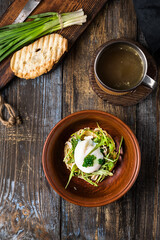Chicken soup with arugula, chicken, poached egg in a deep bowl. On a wooden table are toasted bread, fresh onions, a mug of broth. For the restaurant menu. Top view