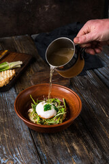 Chicken soup with arugula, chicken, poached egg in a deep bowl. On a wooden table are toasted bread, fresh onions, a mug of broth. A man's hand pours broth into a bowl. Restaurant menu. Close up.