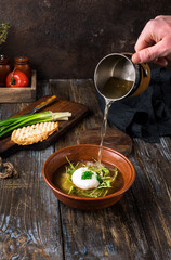 Chicken soup with arugula, chicken, poached egg in a deep bowl. On a wooden table are toasted bread, fresh onions, a mug of broth. A man's hand pours broth into a bowl. Restaurant menu. Close up.