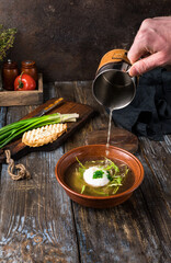 Chicken soup with arugula, chicken, poached egg in a deep bowl. On a wooden table are toasted bread, fresh onions, a mug of broth. A man's hand pours broth into a bowl. Restaurant menu. Close up.