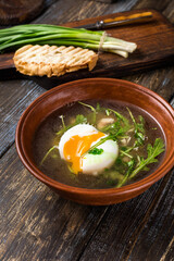 Chicken soup with arugula, chicken, poached egg in a deep bowl. On a wooden table are toasted bread, fresh onions, a mug of broth. For the restaurant menu. Close up.