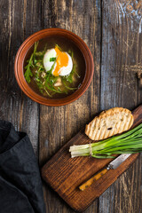 Chicken soup with arugula, chicken, poached egg in a deep bowl. On a wooden table are toasted bread, fresh onions, a mug of broth. For the restaurant menu. Top view