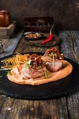 On a serving board, mashed sweet potatoes with bacon rolls with sun-dried tomatoes, microgreens and onion chips. On a wooden table is a box with spices and herbs, a glass of cognac. Restaurant menu