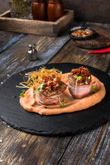 On a serving board, mashed sweet potatoes with bacon rolls with sun-dried tomatoes, microgreens and onion chips. On a wooden table is a box with spices and herbs, a glass of cognac. Restaurant menu