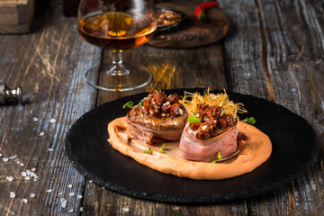 On a serving board, mashed sweet potatoes with bacon rolls with sun-dried tomatoes, microgreens and onion chips. On a wooden table is a box with spices and herbs, a glass of cognac. Restaurant menu
