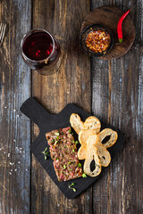 Beef tartare with white bread toast and microgreens on a serving board. On a wooden table a glass of red wine, chili peppers and spices. Menu of a bar, cafe, restaurant. Dish from the chef. Top view.