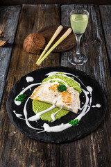 On a round serving board mashed broccoli and vegetables, grilled fish fillet with white cream sauce. On a wooden table a glass of limoncello, spices, lemongrass. Restaurant menu. Close up.