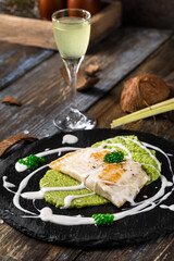 On a round serving board mashed broccoli and vegetables, grilled fish fillet with white cream sauce. On a wooden table a glass of limoncello, spices, lemongrass. Restaurant menu. Close up.