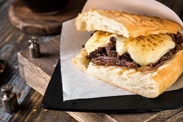Sandwich with beef, cheese and sauce on ciabatta in a paper cover on a wooden table. A glass of dark beer and spices in the composition. Dish for the menu of a restaurant, bar, cafe. Close up