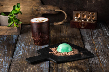 On the serving board is a gourmet dessert green hemisphere with chocolate chips. On a wooden table there is a mug of cappuccino, a sprig of mint in a glass. Close up. Menu for restaurant, cafe, bar.