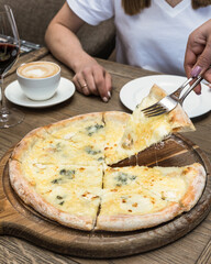 On the table there is a pizza of 4 cheeses cut into pieces, one piece is raised by a female hand. On the wooden table is a cup of cappuccino, a glass of red wine, a plate. 