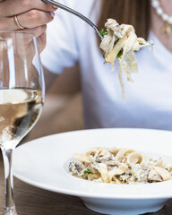Plate of pasta tagliatelle with mushrooms, cheese, herbs and cream sauce close-up. In the foreground is a glass of white wine. The girl picks up the pasta with a fork and lifts it over the plate.