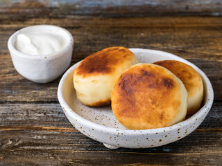 On a white ceramic plate there are 3 curd cheese pancakes and a cup  with sour cream. Wooden background. Close up