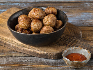 Fried beef meatballs in a dark ceramic bowl. Wooden brown background. Next to a cup of ketchup. Close up.