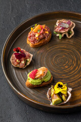 Round brown plate with 5 tapas with beef roast beef, tartare, salmon, chicken and avocado. A dish of haute cuisine. Dark gray textured background. Close up.