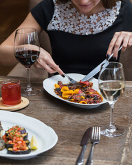 The girl at the table in the restaurant is eating beef carpaccio with a knife and fork. Serving for two, a glass of white and red wine, a mug of red tea, a dish with eggplant in the foreground.