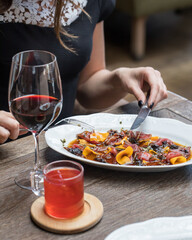 The girl at the table in the restaurant is eating beef carpaccio with a knife and fork. Serving for two, a glass of white and red wine, a mug of red tea, a dish with eggplant in the foreground.