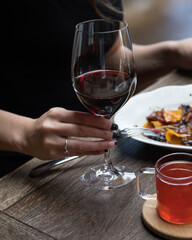 The girl is holding a glass of red wine at the table . In front of her at the table is a plate of food and a mug of red tea. Close up