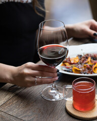 The girl is holding a glass of red wine at the table . In front of her at the table is a plate of food and a mug of red tea. Close up