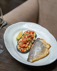 On the table in the restaurant is an oval dish with grilled fish with grilled eggplant cut into slices and vegetables. Close up