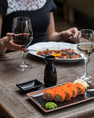 The girl is holding a glass of red wine at the table. A plate of food on the table in front of her. In the foreground is a rectangular plate of sushi, a glass of white wine, soy sauce. Close up.