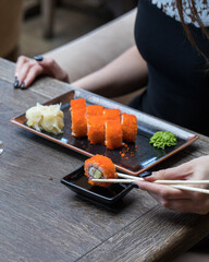 A girl holds a sushi roll with chopsticks over soy sauce. On the wooden table in front of her is a rectangular plate with sushi, wasabi and ginger. Close up