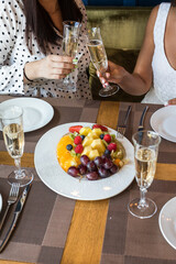 Two girls at a table in a restaurant clink glasses of champagne. The table is served with fruit and plates.