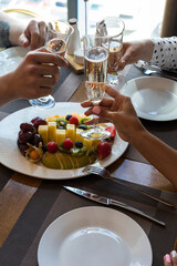 2 girls and 1 guy at a table in a restaurant clink glasses of champagne. The table is served with fruit and plates.