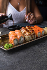 Rectangular plate with sushi rolls, wasabi and ginger, sushi sticks. In the background, a girl in white pours soy sauce into a saucepan. Close up.